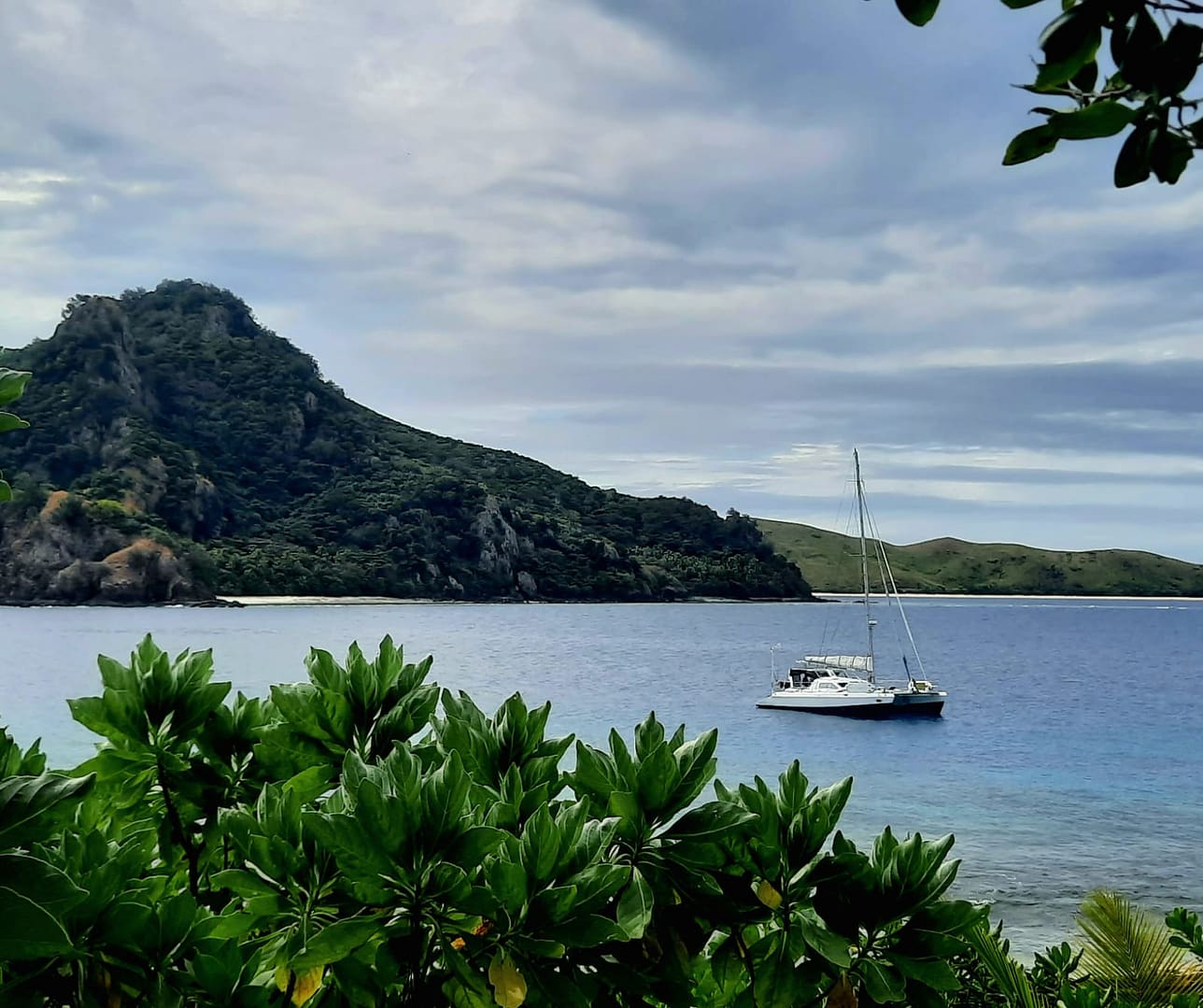 A scenic view of a boat anchored in a calm bay with lush green foliage in the foreground, a mountain on the left, and rolling hills in the background under a cloudy sky.