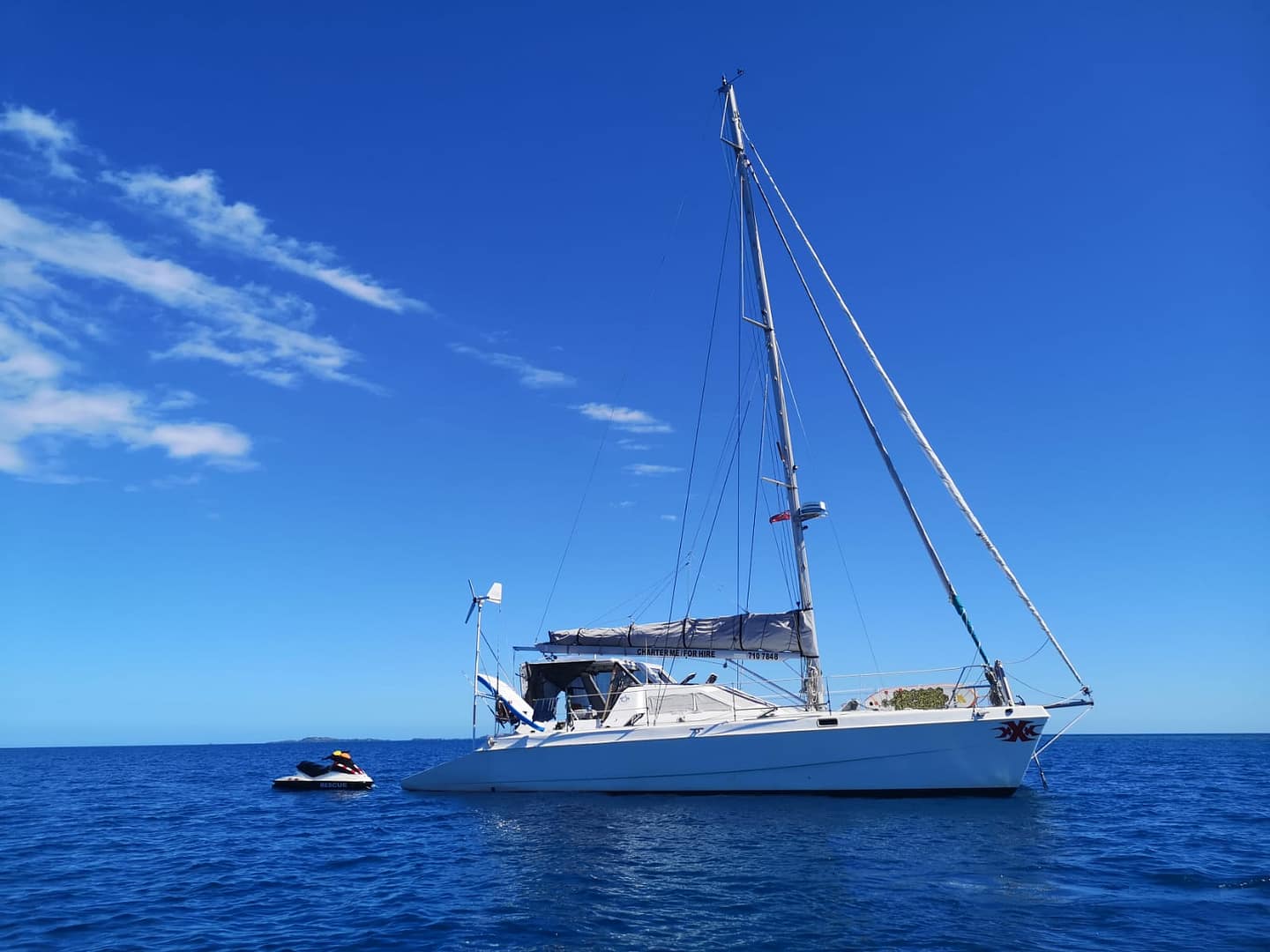 White sailboat with mast and rigging, anchored on calm blue ocean waters under a clear blue sky with scattered clouds, accompanied by a small black and red rescue jet ski tethered to its side.