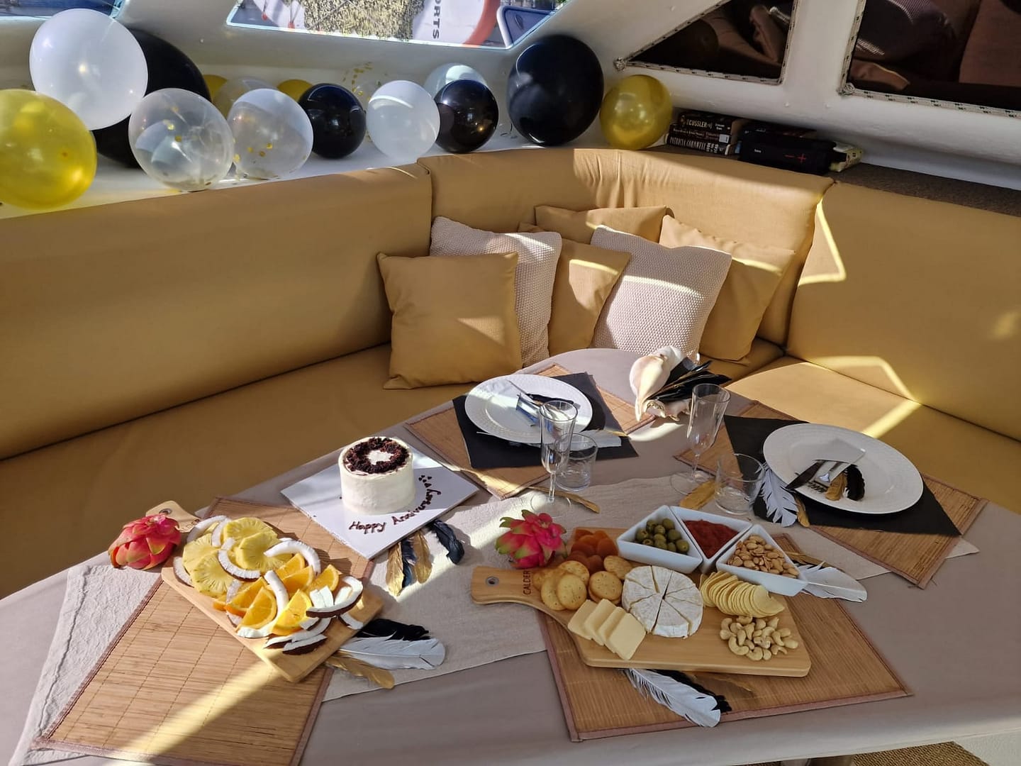 A table set for a celebration with a cheese platter, fruit, crackers, a small cake with "Happy Anniversary" written on it, and empty glasses. The background has a yellow cushioned seating area, and balloons in black, white, and yellow are placed along the window ledge.