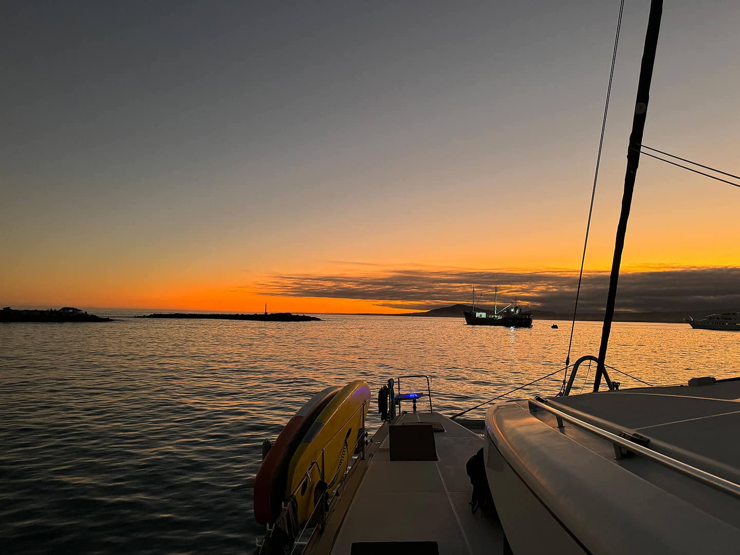 A view from the deck of a boat at sunset, showing calm waters, with other boats and a lighthouse in the distance under a colorful sky.