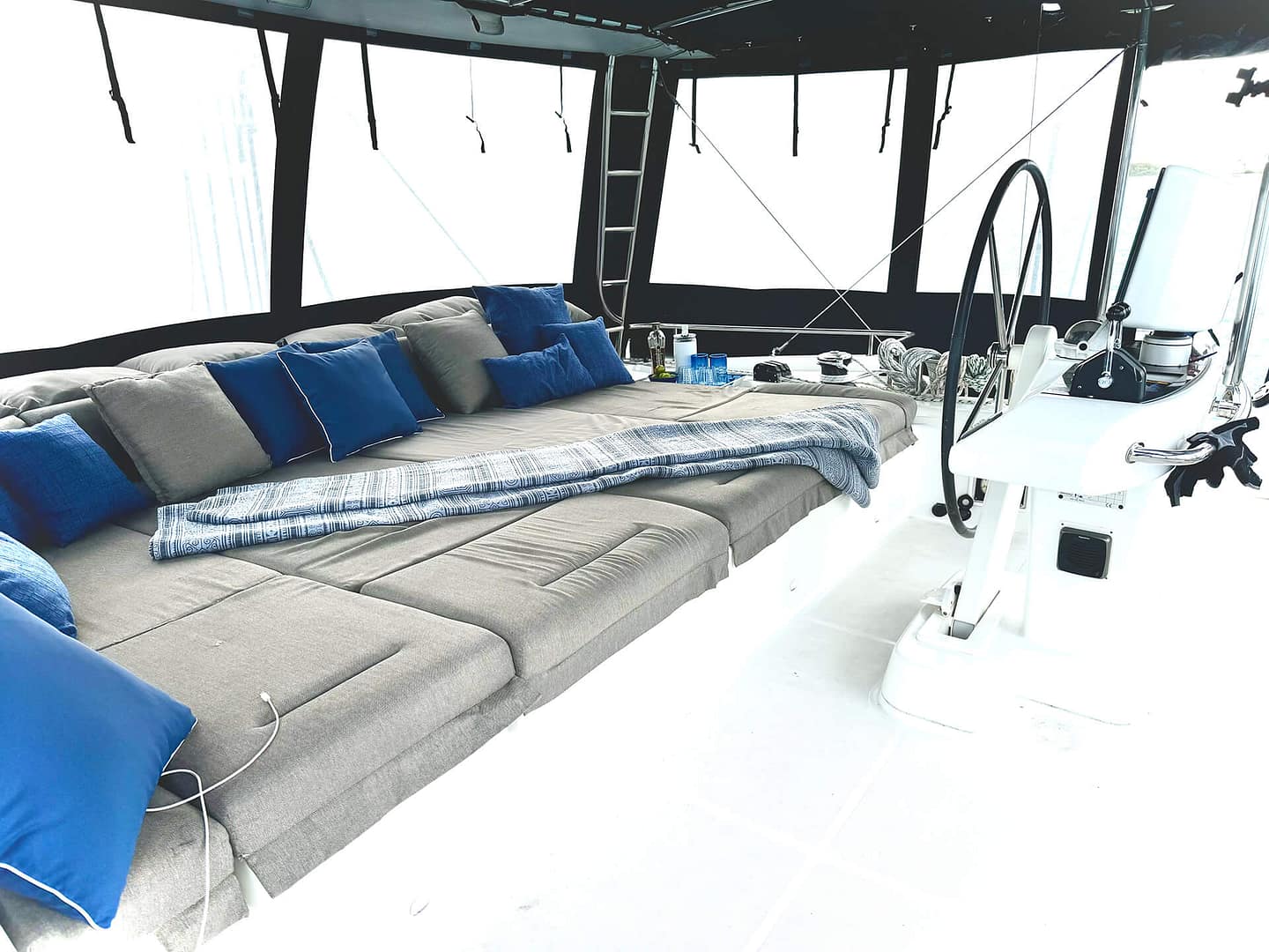 A spacious, cushioned lounge area on a boat with numerous blue and beige pillows, overlooking large windows and surrounded by nautical equipment.