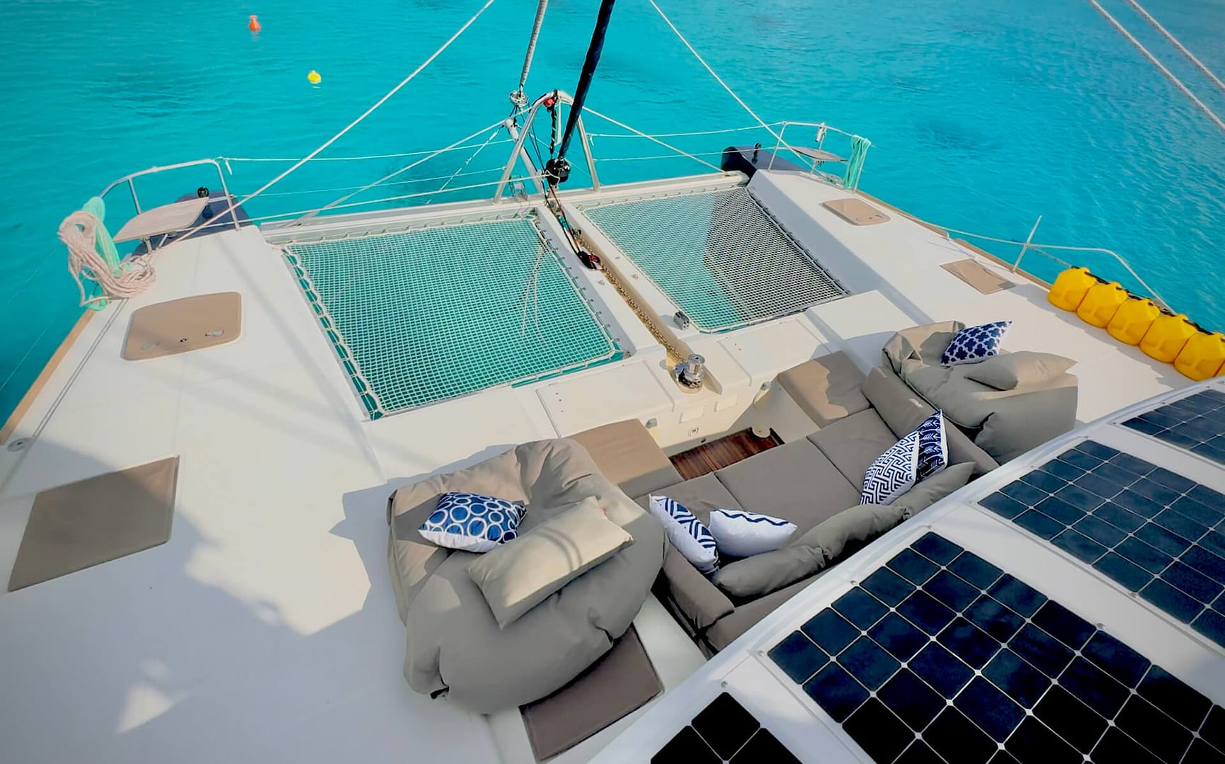 A yacht deck with outdoor seating, cushions, and pillows, featuring solar panels and a netted trampoline, floating on calm blue water.