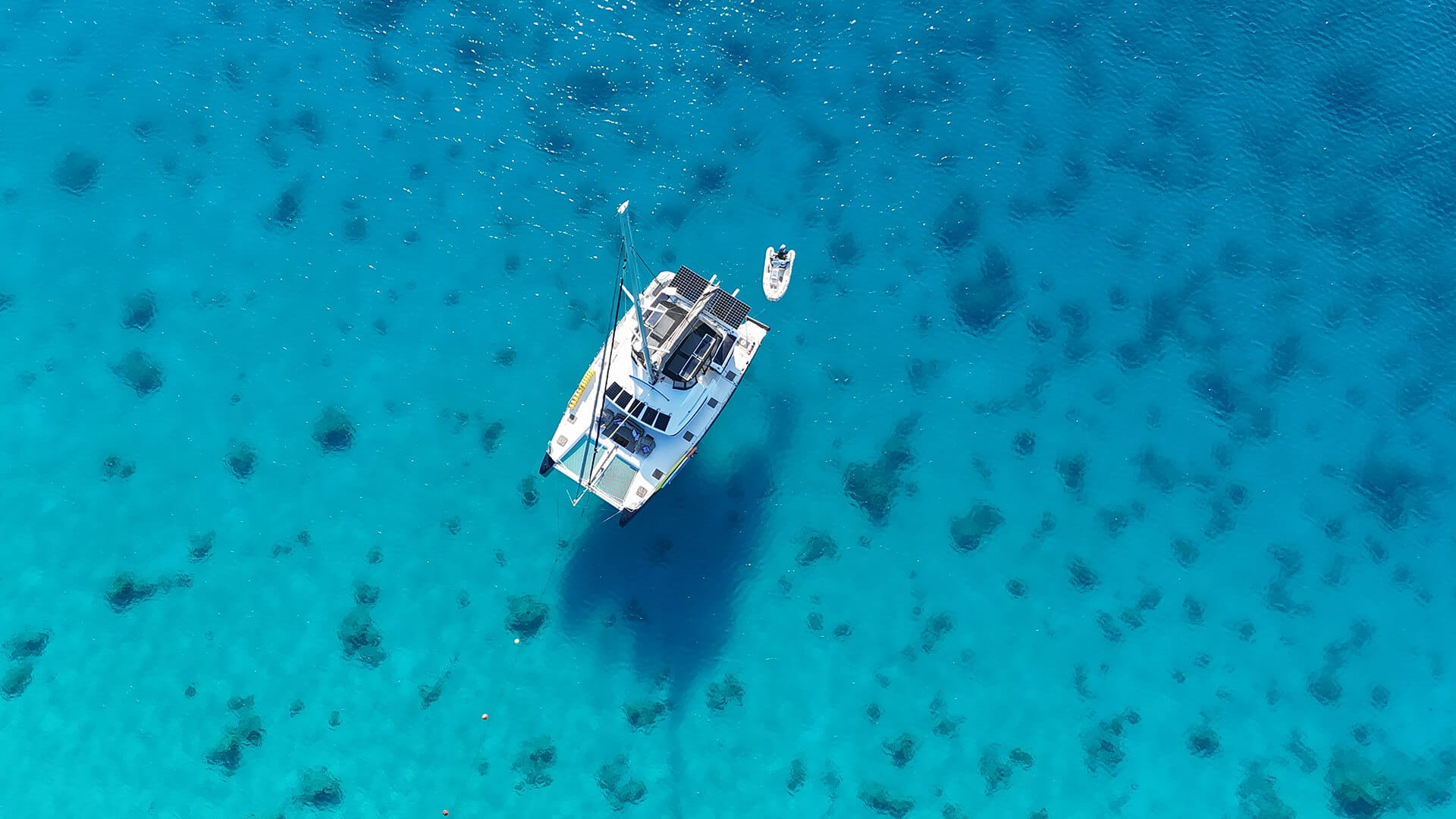 Aerial view of a white yacht with solar panels floating on clear blue water with visible dark patches and gentle ripples.