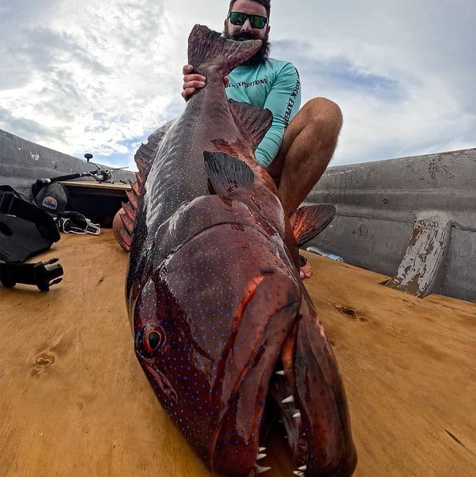 A man with sunglasses and a beard crouches behind a large fish on a wooden surface, with fishing gear and a cloudy sky in the background.