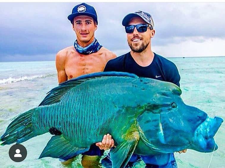 Two men standing in shallow ocean water holding a large, brightly colored fish together, with overcast sky in the background.
